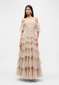 Γυναίκα που φοράει μια μπεζ τουαλέτα με βαθμίδες, βολάν και διαφάνεια, με υφή και σχέδια, στέκεται μπροστά σε ένα απλό λευκό φόντο.