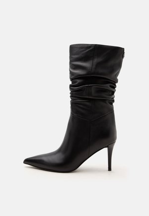 Botte en cuir noire mi-mollet avec bout pointu, tige relâchée et talon haut et fin sur fond blanc.