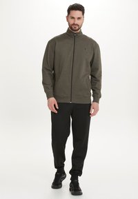 Mörkgrön zip-up tröja med hög krage, ribbade mudd och fåll. I kombination med svarta byxor och svarta sneakers. Enkel logotypdetalj.