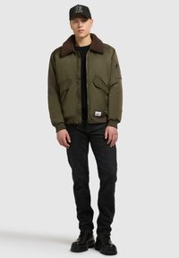 Olivgrüne Bomberjacke mit einem braunen Kunstfellkragen, Reißverschluss, zwei Fronttaschen, getragen mit schwarzen Jeans und Stiefeln, vor einem neutralen Hintergrund.