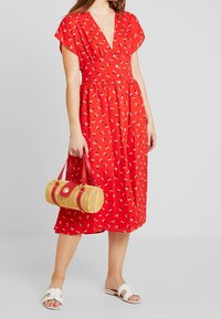 Vestido floral rojo con un escote en V profundo y mangas cortas, ceñido en la cintura. La modelo lleva un bolso redondo de mimbre con acentos rojos y sandalias blancas.