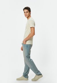Lys beige ribbet t-shirt parret med falmede blå jeans og beige sneakers; modellen står sidelæns mod en ensfarvet hvid baggrund.