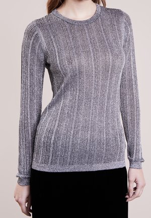Mujer con un suéter ajustado de manga larga, plateado metálico y de tejido acanalado, con cuello redondo, sobre un fondo beige liso.