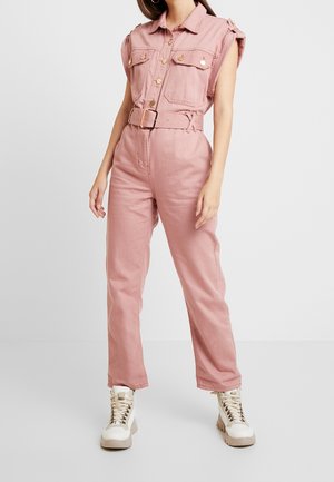 Frau trägt einen ärmellosen, staubrosa Jumpsuit mit Gürtel und goldenen Knöpfen sowie weiße High-Top-Sneaker und steht vor einem schlichten Hintergrund.