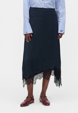 Personne portant une jupe asymétrique bleu marine avec une bordure en dentelle noire, une chemise à rayures bleues et des chaussures Mary Jane rouge foncé, debout sur fond blanc.
