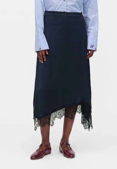 Personne portant une jupe asymétrique bleu marine avec une bordure en dentelle noire, une chemise à rayures bleues et des chaussures Mary Jane rouge foncé, debout sur fond blanc.