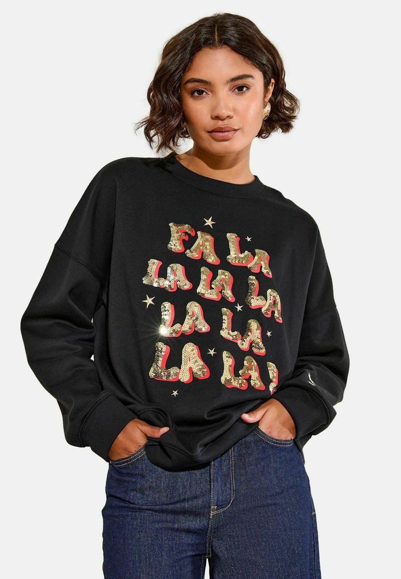 Melns sporta krekls ar brīvu piegriezumu, kurā ir zelta un sarkanu pailgu tekstu "FALALA" un mazu zvaigznīšu akcentus. Ribotas aproces un apkakle.