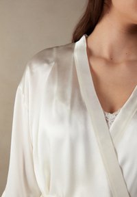 Bílý saténový župan s jemným leskem, s kontrastní krajkovou vsadkou ve výstřihu a páskem pro zapínání, který vyniká hladkým povrchem.