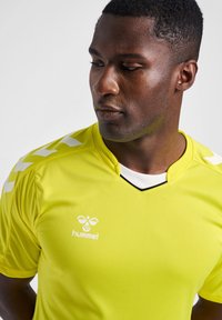 Geel sportshirt van gladde stof, met een v-hals, witte accenten op de schouders en een logo op de borst.