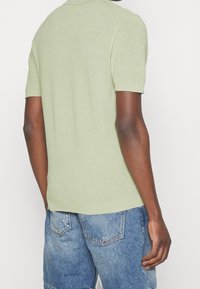 Koszulka polo z krótkim rękawem wykonana z jasnozielonego dzianego materiału, charakteryzująca się fakturowaną powierzchnią i klasycznym kołnierzykiem. Noszona z niebieskimi szortami z denimu.