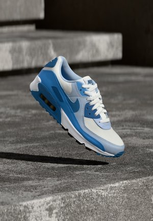 Modrá a biela teniska Nike visí vo vzduchu nad betónovými schodmi, zvýrazňujúc svoj dizajn a textúrované detaily podrážky.