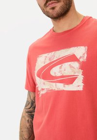 Koralfarvet bomulds T-shirt med et stort hvidt grafisk logo på en tekstureret baggrund. Rund halsudskæring og korte ærmer.
