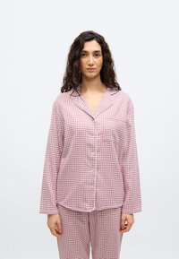 Γυναίκα με μακριά σγουρά σκούρα μαλλιά, φορώντας ροζ και λευκό πιε ντε πουλ κουμπωτό πιτζάμα σετ, στέκεται με τα χέρια χαλαρά σε λευκό απλό φόντο.