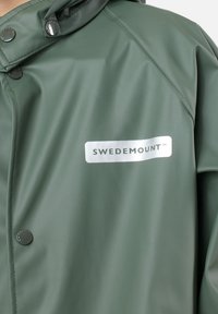 Giacca impermeabile verde con una texture liscia, etichetta di branding argentata e bottoni a pressione sul davanti per la chiusura.