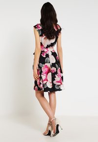 Kvinna som står med ryggen vänd, iklädd en svart knälång blommig klänning och beige högklackade sandaler mot en enfärgad bakgrund.