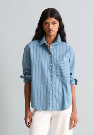 Junge Frau mit langen dunklen Haaren trägt ein blaues, vertikal gestreiftes Hemd mit Knöpfen und hochgekrempelten Ärmeln sowie eine weiße Hose vor einfachem Hintergrund.