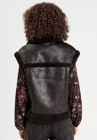 Gilet in pelle nera con bordo in pelliccia, superficie testurizzata e senza maniche, abbinato a una camicia a maniche lunghe con un motivo colorato sottostante.