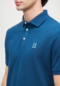 Blue polo shirt made of textured cotton. Features a two-button placket and an embroidered white logo on the left chest. Short sleeves.