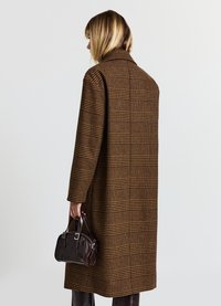 Brauner Karomantel mit strukturierter Form, breitem Kragen und langen Ärmeln. Dunkle Handtasche mit silbernen Beschlägen, in der rechten Hand gehalten.