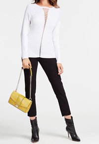 White long-sleeve top with a v-shaped cutout featuring lace-up detailing, paired with black fitted pants and black ankle boots. Holds a yellow handbag.