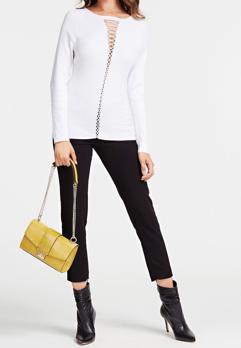 White long-sleeve top with a v-shaped cutout featuring lace-up detailing, paired with black fitted pants and black ankle boots. Holds a yellow handbag.