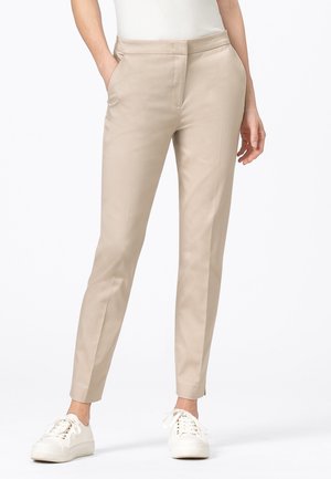 Beige slim-fit broek gedragen met witte vetersneakers en een wit ingestopt topje, handen in de zakken, staand tegen een witte achtergrond.