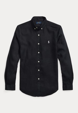 Άνδρας με μαύρο μακρυμάνικο πουκάμισο, λευκά σορτς με ψηλή μέση, μαύρα γυαλιά ηλίου και μαύρα παπούτσια χωρίς κορδόνια σε απλό φόντο.