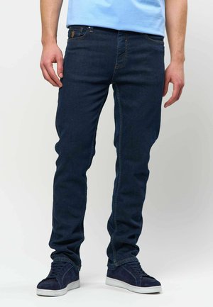 Mężczyzna w ciemnoniebieskich dopasowanych dżinsach, jasnoniebieskiej koszuli i granatowych tenisówkach stoi na jednolitym białym tle.