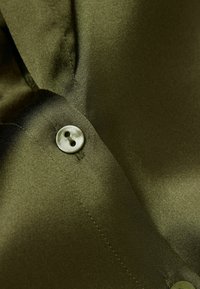 Tessuto di raso verde con una texture liscia, caratterizzato da un bottone rotondo e dettagli di cucitura. Vista ravvicinata di una sezione del colletto di una camicia.