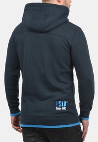 Donkerblauwe hoodie met een gladde textuur, capuchon en contrasterende turquoise accenten op de mouwen en onderrand. Bevat een logo en tekst op de achterkant.