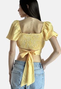 Cropped top giallo a scacchi con maniche a sbuffo, corpetto arricciato e un nodo sul retro. Il tessuto presenta un motivo a quadri e una texture morbida.