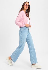 Roze satijnen bomberjack met elastische manchetten, gecombineerd met lichtblauwe wijde jeans en crèmekleurige sneakers, met een trekkoord in de taille.