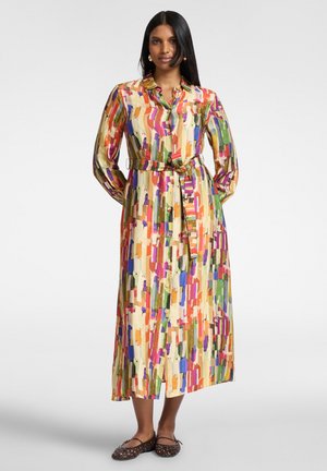 Vrouw staat en draagt een kleurrijke midi-jurk met lange mouwen en knopen, met een abstract patroon van verticale penseelstreken en een ceintuur die om de taille is gestrikt, bruine platte schoenen.