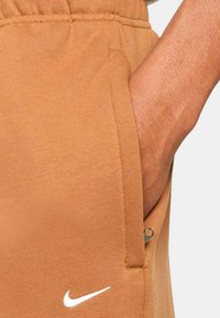 Nike joggers dalam warna coklat hangat, dilengkapi dengan saku samping yang memiliki ritsleting dan logo putih kecil dekat penghematan, terbuat dari bahan lembut.