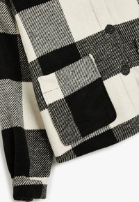 Zwart-wit geblokte jacket met een gestructureerde stof, enkele klepzak en zwarte knopen, met opvallende contrasterende patronen.