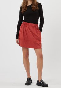 Jupe rouge texturée avec une taille élastique et des poches latérales. Associée à un haut noir à manches longues et des mocassins noirs épais.