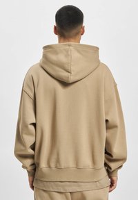 Hoodie oversize beige en tissu doux. Présente une grande capuche, des épaules tombantes et des poignets et un ourlet côtelés pour le confort.