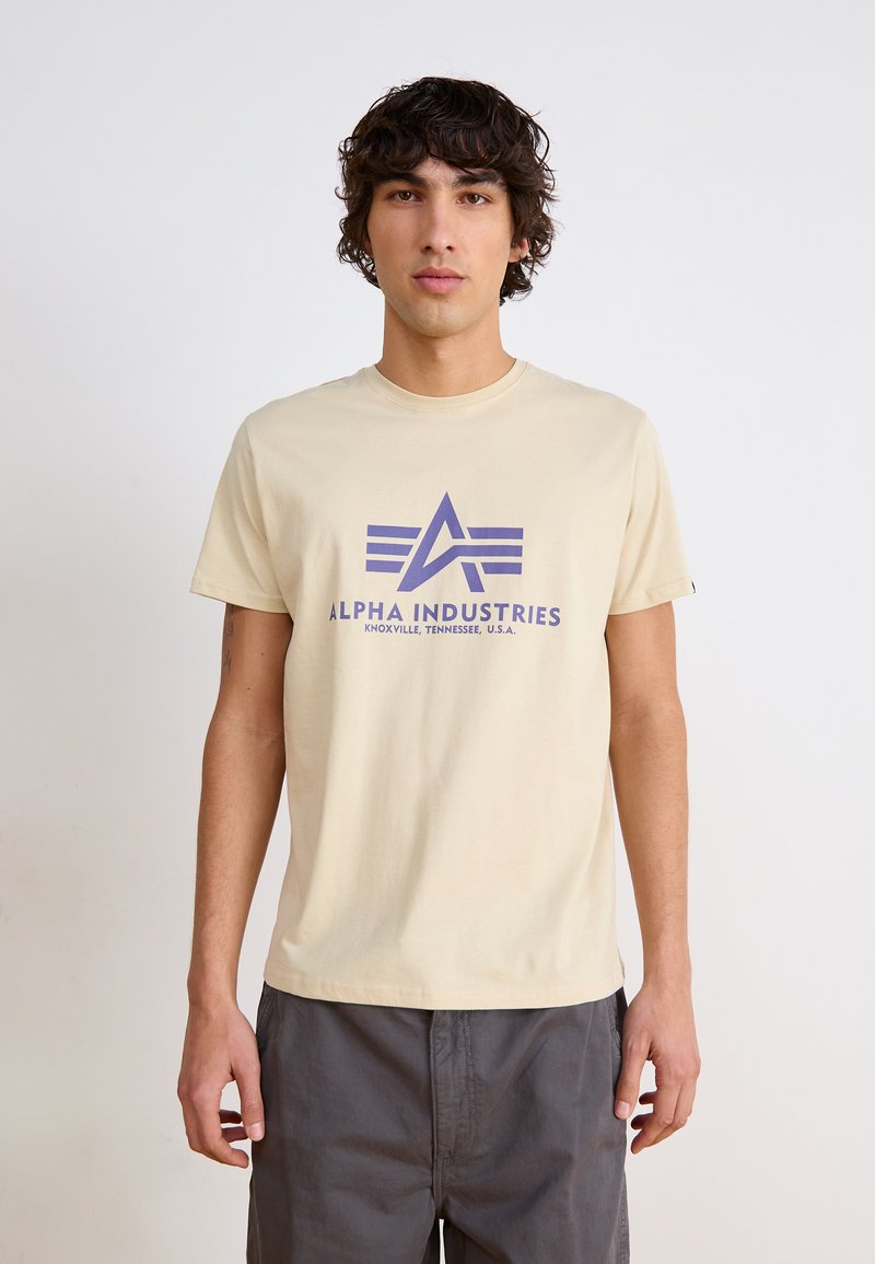 Beige bomulds T-shirt med et lilla grafisk logo af et A med tre vandrette striber. Korte ærmer og rund halsudskæring, afslappet pasform.