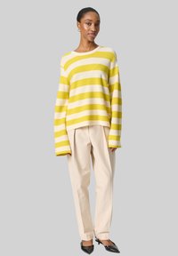 Pull rayé jaune et crème, coupe ample avec des manches larges; associé à un pantalon beige clair et des chaussures noires à talons pointus.