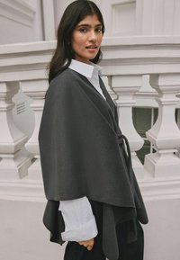 Mörkgrå, ullponcho med knytning, draperad design, lagerkanter och en kontrasterande vit skjorta under.