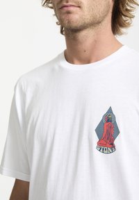 Hvid bomulds t-shirt med et grafisk print på venstre bryst, med røde, blå og sorte accenter, der forestiller en siddende figur og geometriske former.