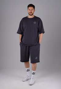 Dunkelgrauer, übergroßer Sweatshirt und passende Shorts, aus weichem Material gefertigt. Verfügt über kurze Ärmel und ein Logo auf der linken Seite. Weiße Sneaker und Socken.