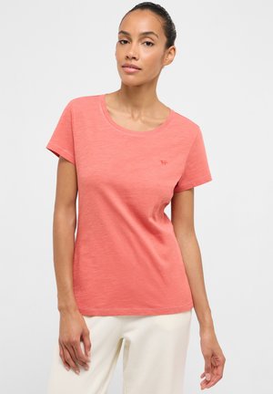 Frau mit zurückgebundenen Haaren, die ein korallenfarbenes Kurzarmshirt und helle Hosen trägt, steht vor einem einfarbigen hellen Hintergrund.