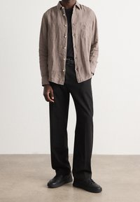 Jasnobrązowa lniana koszula zapinana na guziki z jedną kieszenią na piersi, zestawiona z czarnymi spodniami i czarnymi trampkami. Prosty, luźny krój.