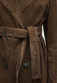 Trench-coat en daim marron avec des boutons noirs et une ceinture assortie nouée à la taille, montrant des coutures détaillées et une texture apparente.