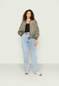 Ελαιόχρωμο bomber jacket με χαλαρή εφαρμογή, ριγωτούς μανίκους και φερμουάρ μπροστά; σε συνδυασμό με ανοιχτό μπλε τζιν ίσιας γραμμής και αθλητικά παπούτσια.
