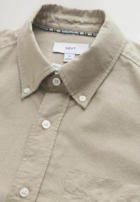 Chemise beige à boutons avec col, boutons blancs, et une étiquette visible indiquant la taille moyenne et la marque "Next".