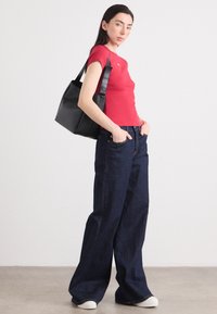 Une femme se tient dans une pose décontractée, portant un haut rouge ajusté et un jean en denim à jambes larges, tenant un sac fourre-tout noir, sur un fond uni.