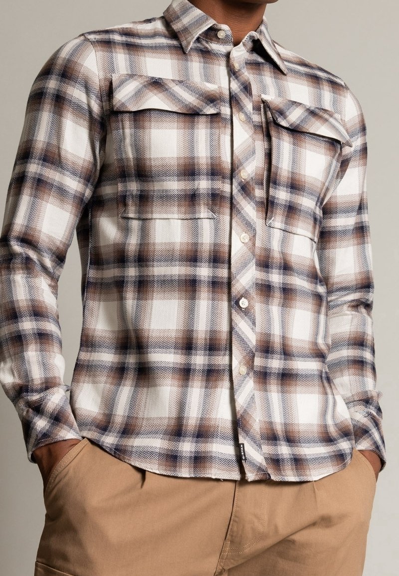 Homme portant une chemise à carreaux marron, blanche et grise avec deux poches poitrine et un pantalon beige, les mains dans les poches.