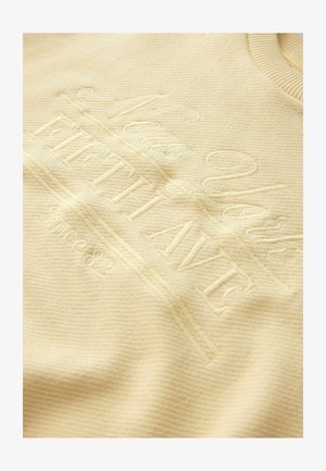 Beige tekstureret stof med hævet broderetekst, der lyder "New York FAITH ALIVE Since 82" nær en ribbet halsudskæring.